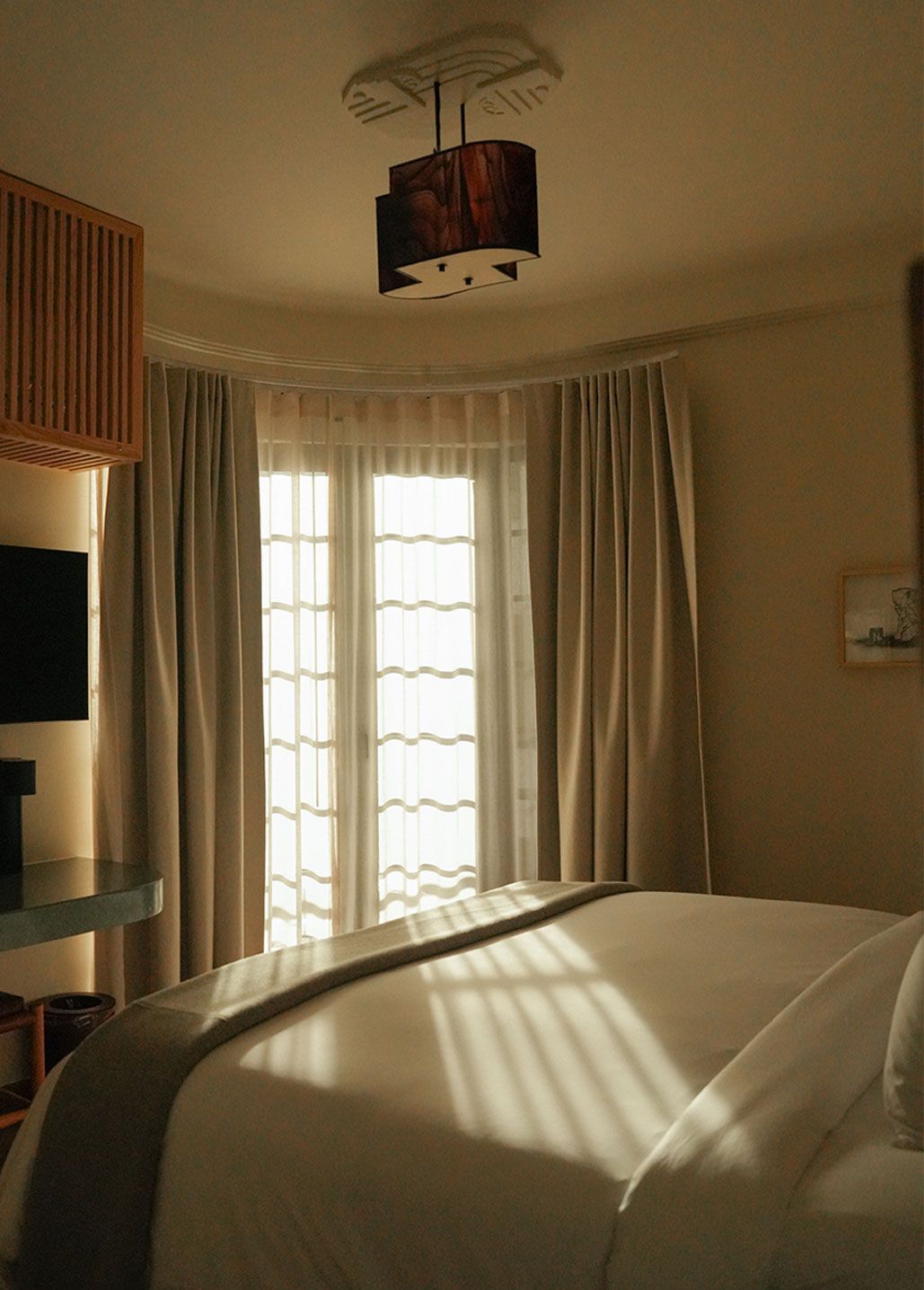 Dormitorio con cama, ventana con cortinas y lámpara de madera. La luz del sol entra a raudales.