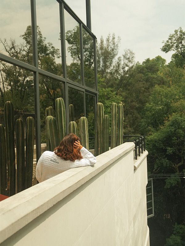 Mujer en un balcón, contemplando cactus y árboles altos. Cielo nublado, barandilla blanca y ventana de cristal.