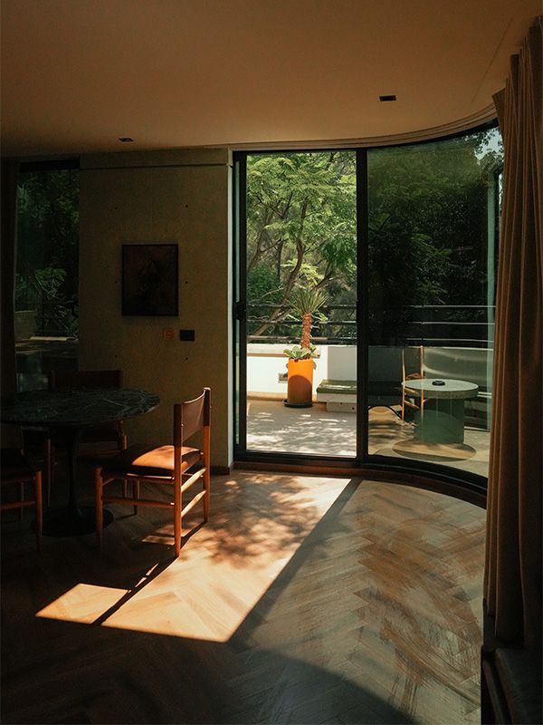 Interior iluminado por el sol con pisos de madera, pared de vidrio curva que conduce a un balcón con una mesa y una exuberante vegetación.