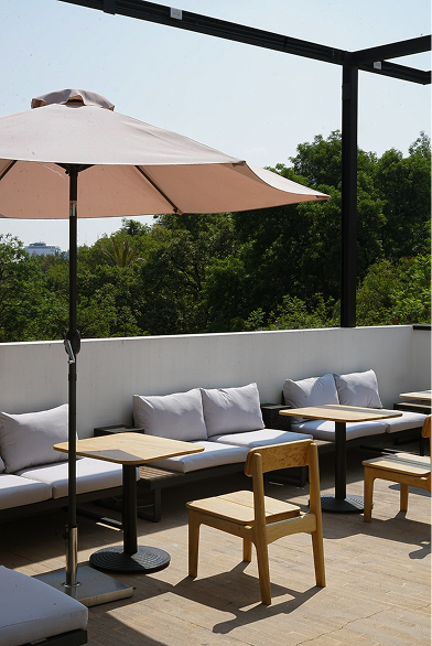Patio al aire libre con sofás gris claro, mesas y sillas de madera y una sombrilla color melocotón.