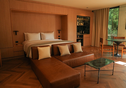Dormitorio con cama, sofá de cuero, mesa pequeña y barra. Revestimiento y suelos de madera color marrón claro.