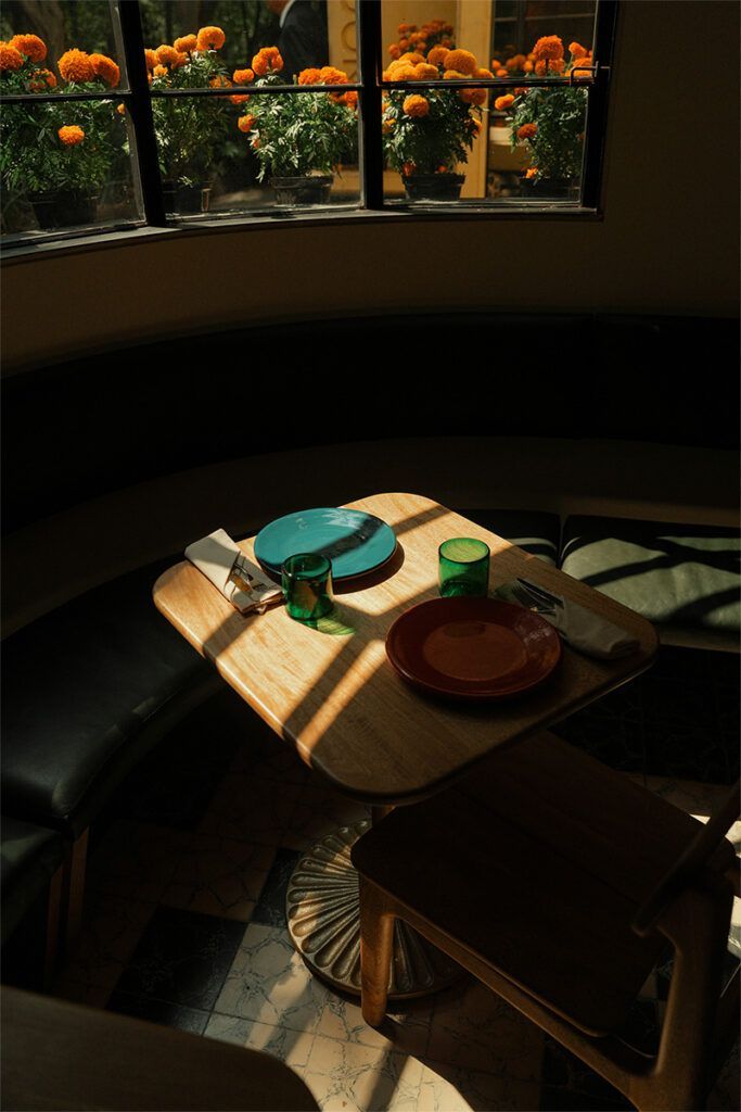 Mesa puesta para dos junto a una ventana soleada, con platos coloridos y flores naranjas en el exterior.