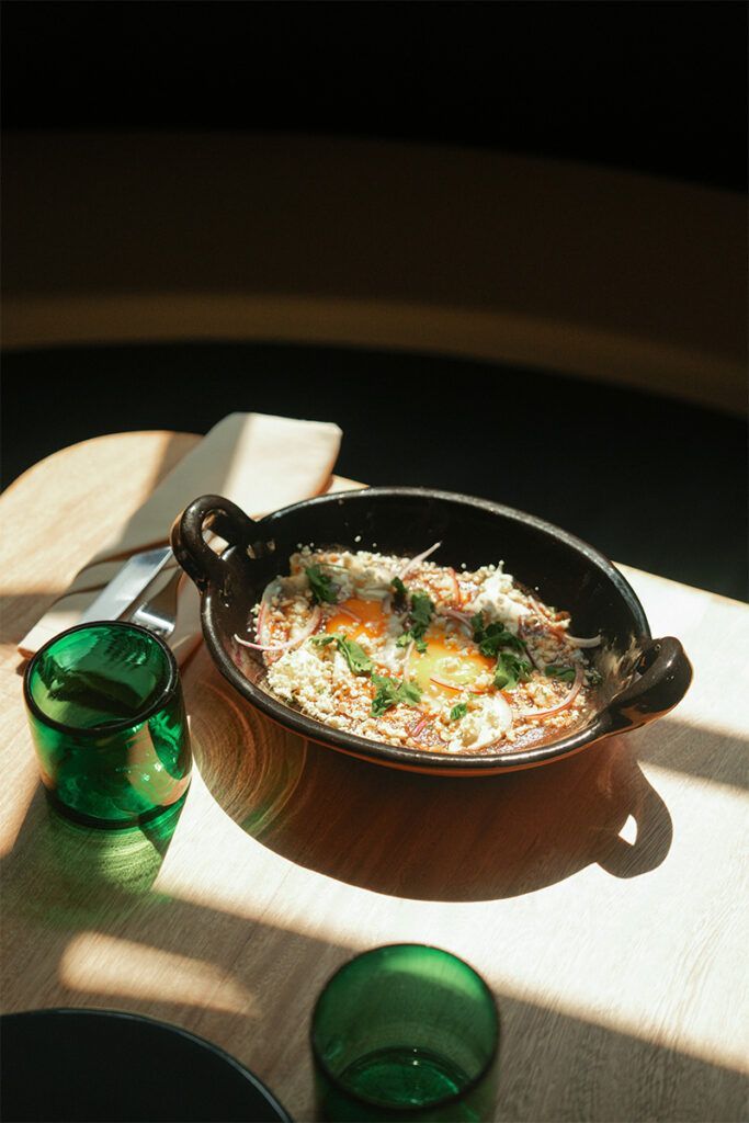 Plato horneado con huevo, queso y hierbas en una sartén negra sobre una mesa de madera con vasos verdes.