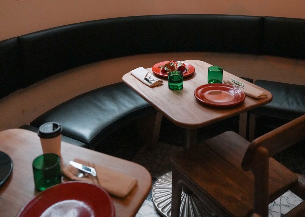 Interior de restaurante con mesas de madera, platos rojos, vasos verdes y una cabina negra curvada.