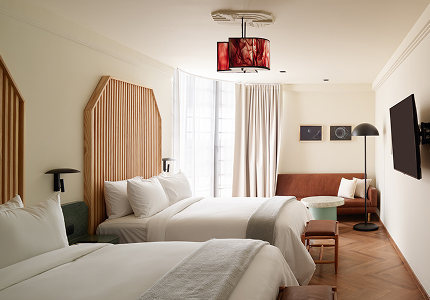 Habitación de hotel con dos camas, sofá, televisor y una lámpara única. Tonos crema y marrón claro.