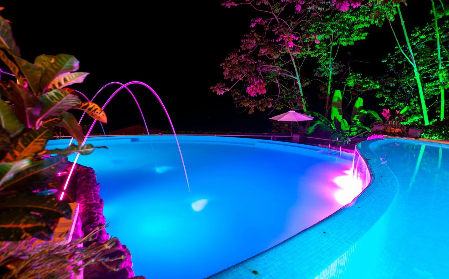 A large swimming pool is lit up at night with purple and green lights.