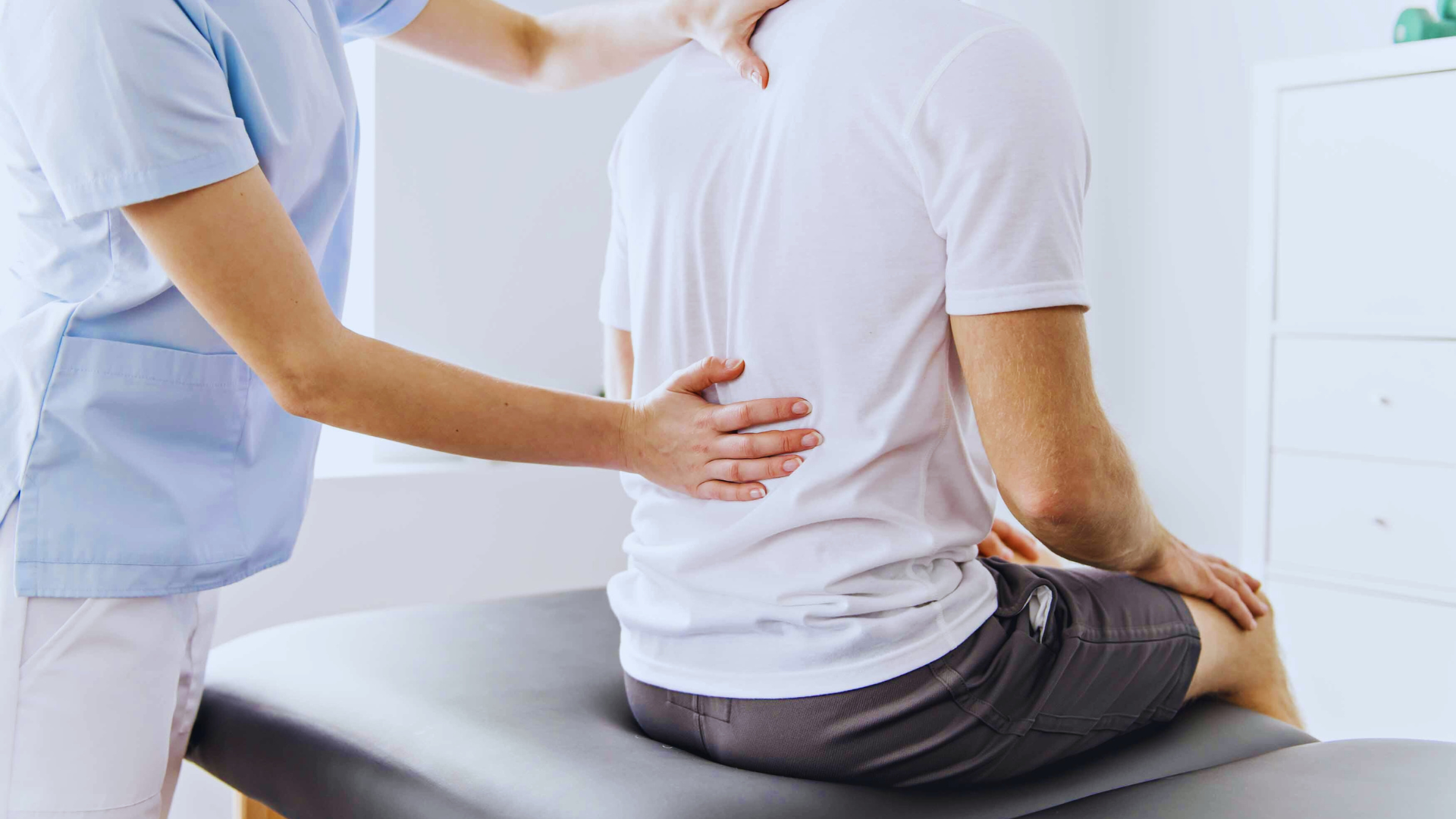 Physiotherapist examining a seated patient's back in a light-filled treatment room.