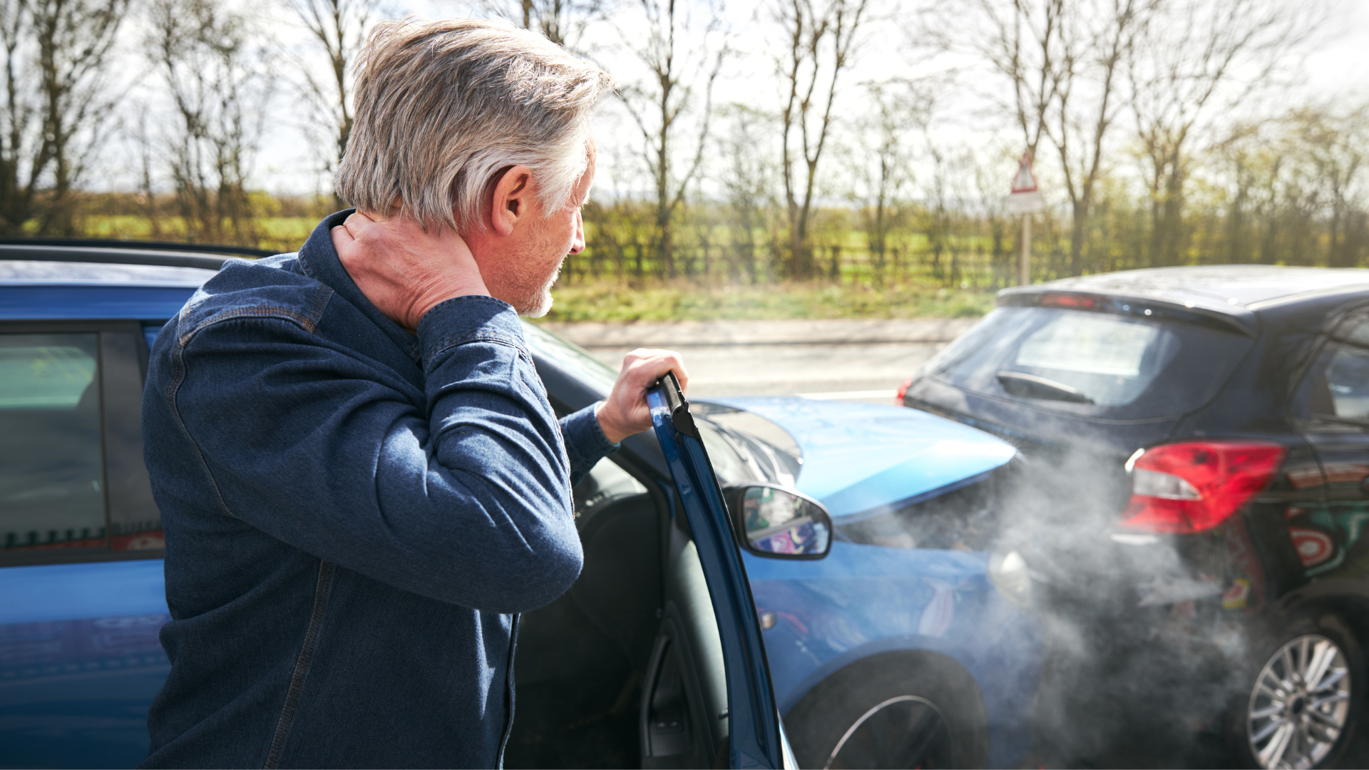 Man holding his neck next to a damaged blue car after a collision with a black car. Steam rising.