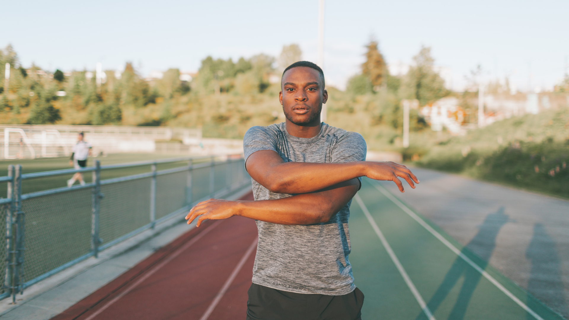 Man stretching on a track, arms crossed, green field in background.