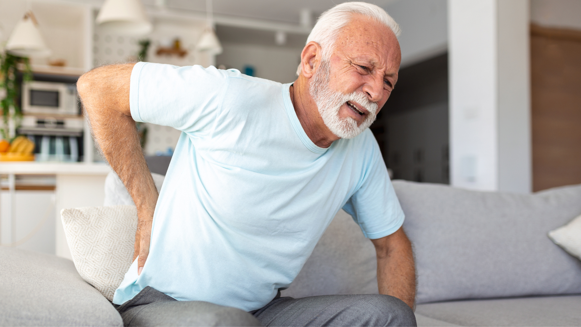 Older man with gray hair, grimacing in pain, holding his lower back while sitting on a couch.