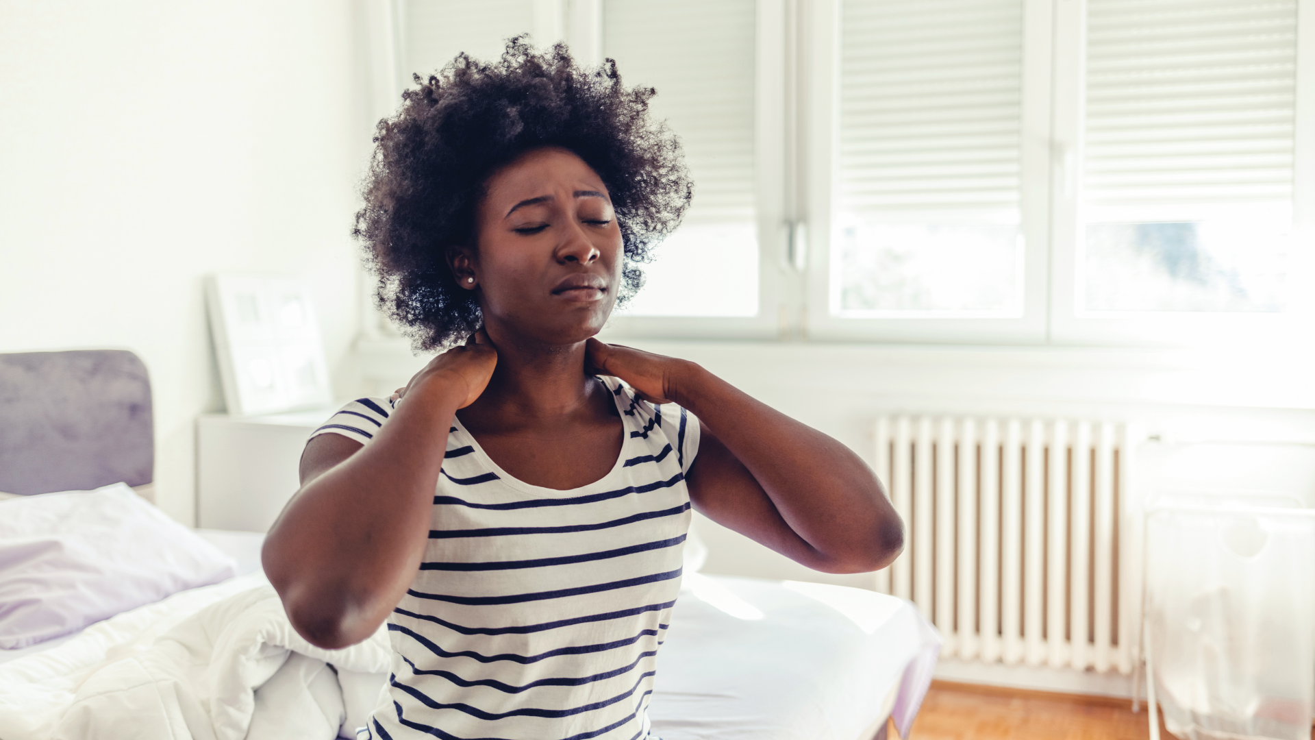 Woman in striped shirt holding her neck in pain, near a bed and window.