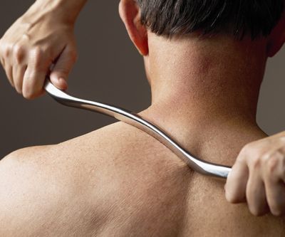 Person receiving a back massage with a curved metal tool on their upper back and neck.