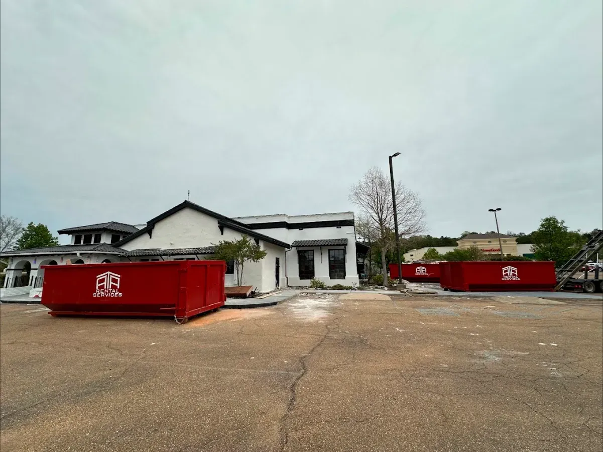 There are two red dumpsters in front of a white house.