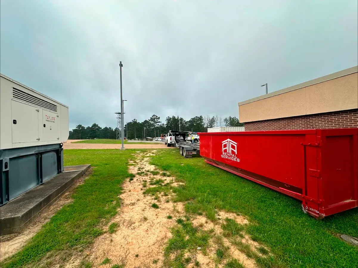 A red dumpster is sitting in the grass next to a building.