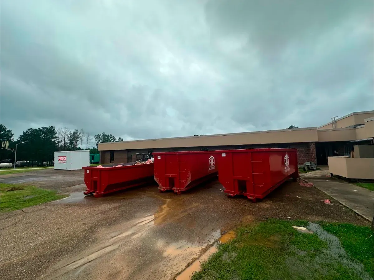 Three red dumpsters are parked in front of a building on a cloudy day.