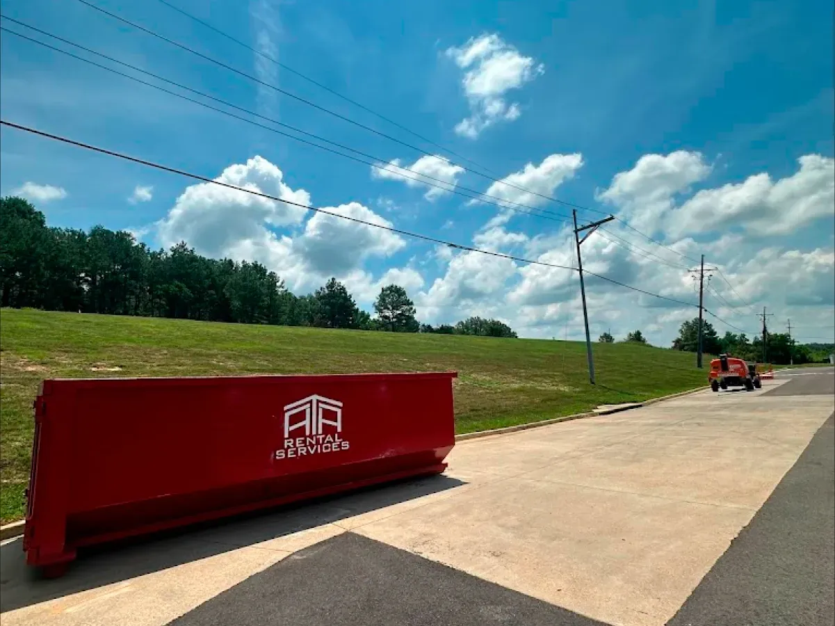 A red dumpster is parked on the side of the road next to a grassy field.