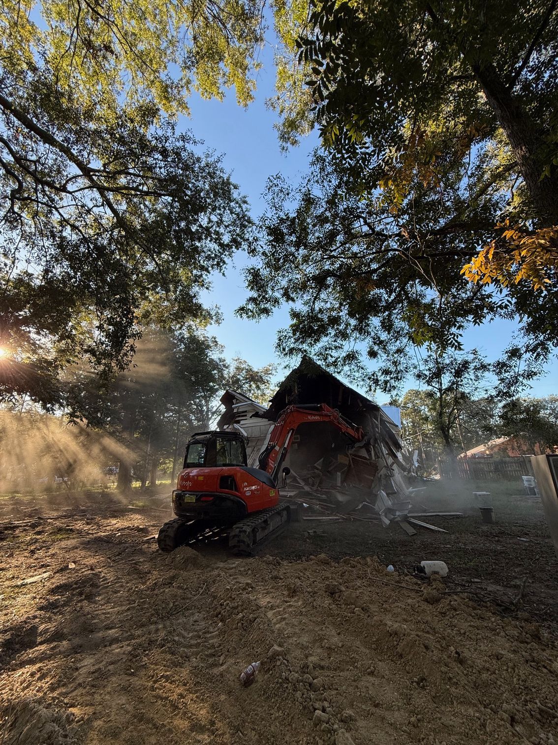 An excavator is demolishing a house in a field surrounded by trees.