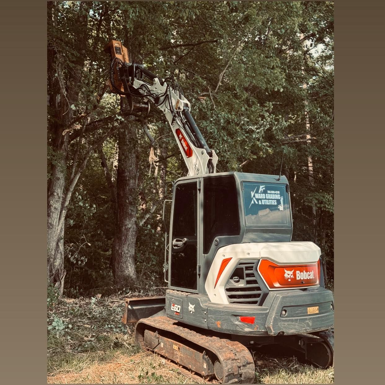 Excavator clearing brush in a wooded area, with its boom raised among trees.