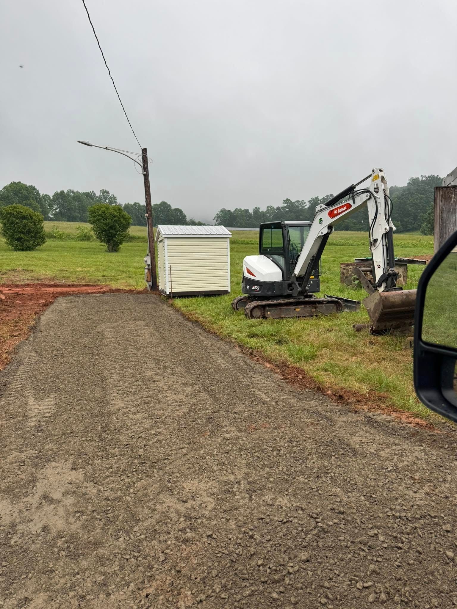 Gravel driveway beside a white container, with a small excavator on a grassy roadside under cloudy skies.
