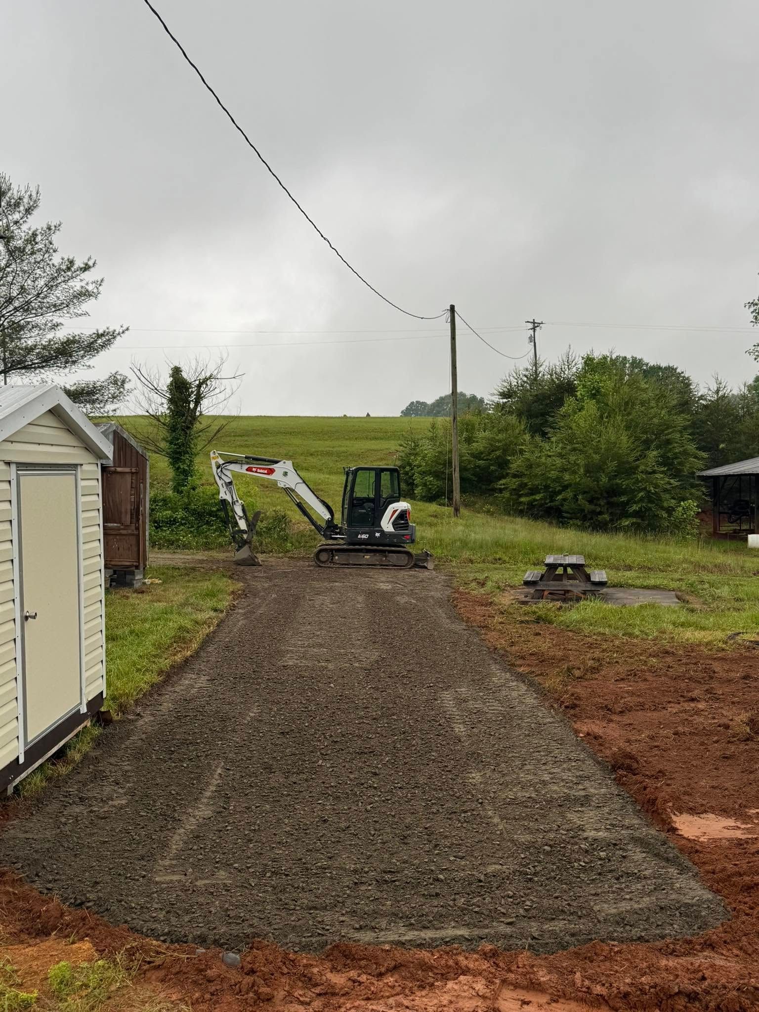 Gravel driveway under construction with a mini excavator and utility poles on a cloudy day