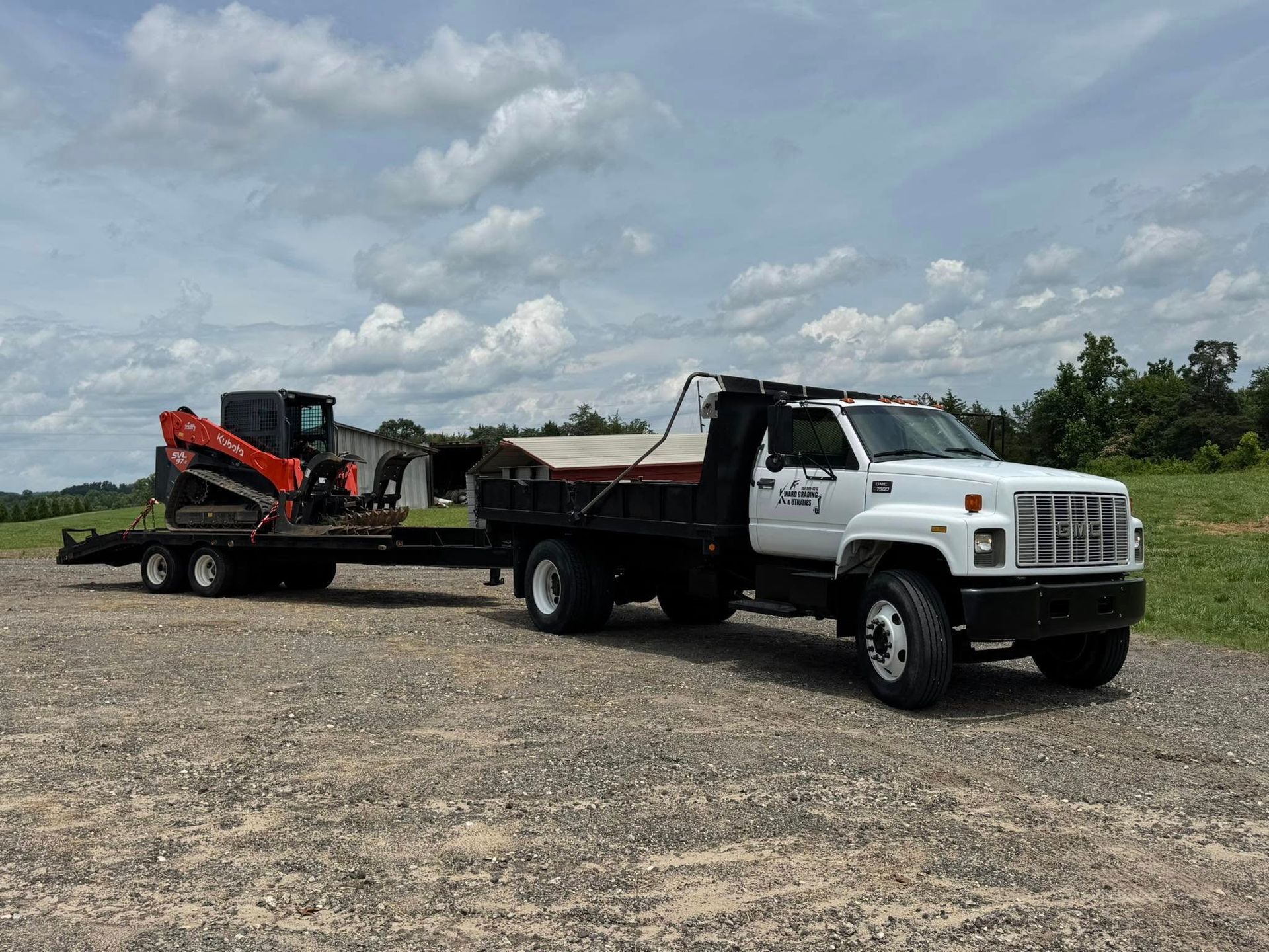 White dump truck towing a red skid steer on a flatbed trailer across a gravel lot under a cloudy sky