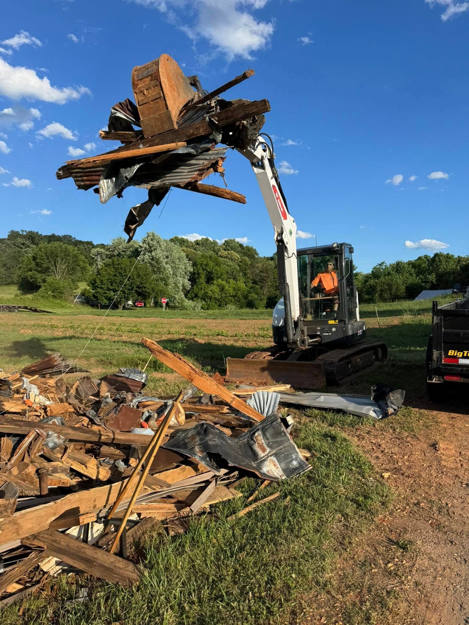 Excavator lifting wooden debris and roof remains in a grassy yard under a blue sky