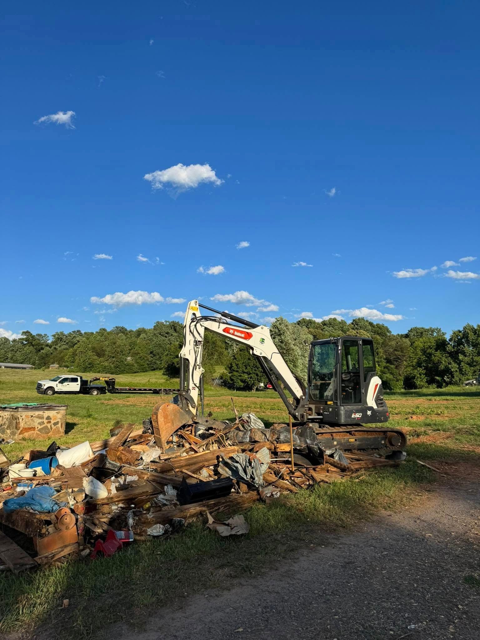 Excavator in a rural field beside a pile of debris under a bright blue sky