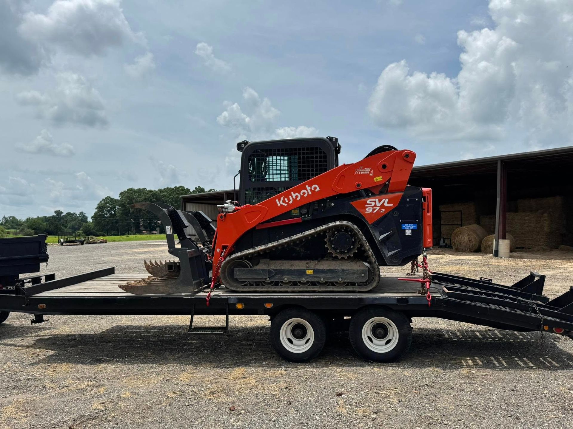 Orange Kubota skid steer on a flatbed trailer in a farm yard under cloudy skies