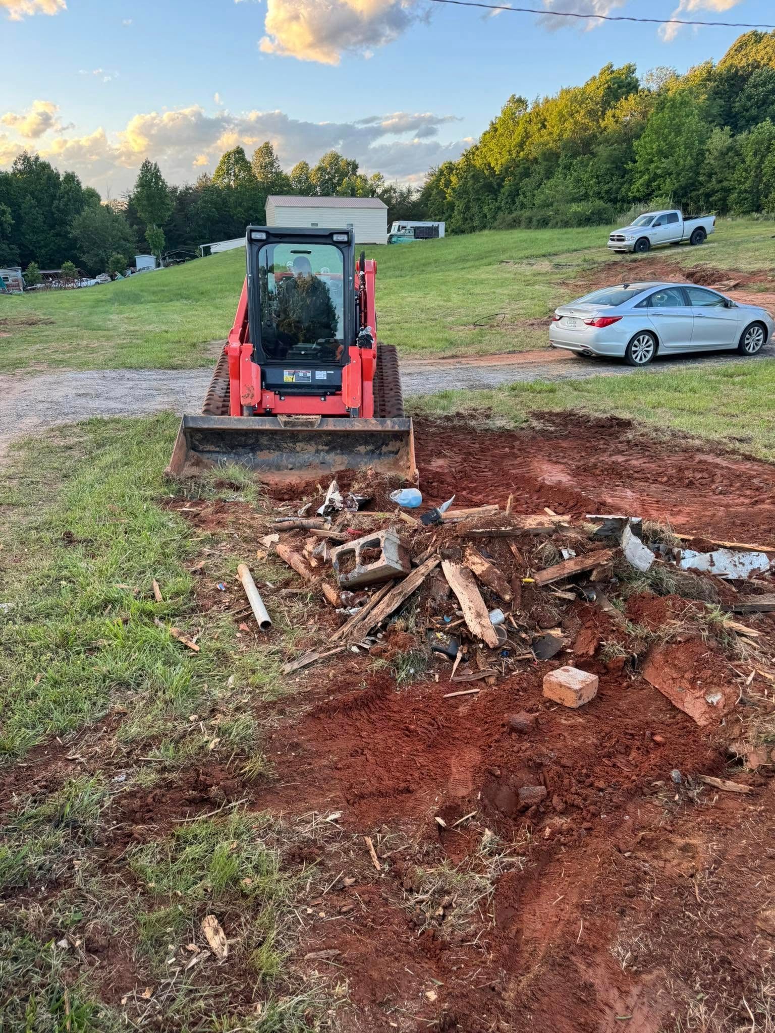 Red skid steer clearing debris from a muddy lot with a car and wooded hillside nearby