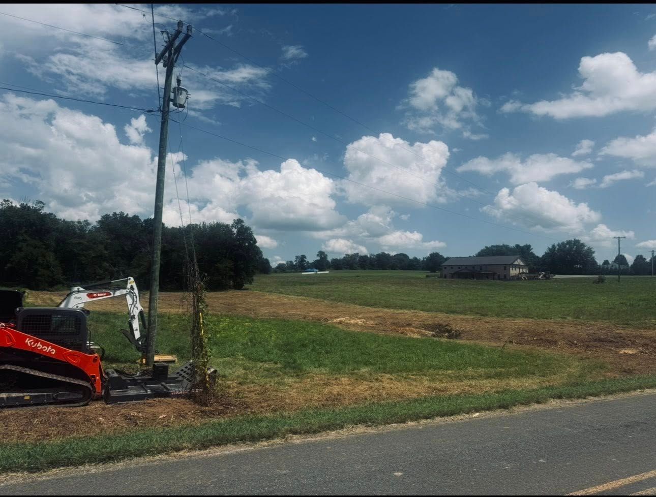 Freshly graded red dirt lot beside a house and wooded edge under a clear blue sky