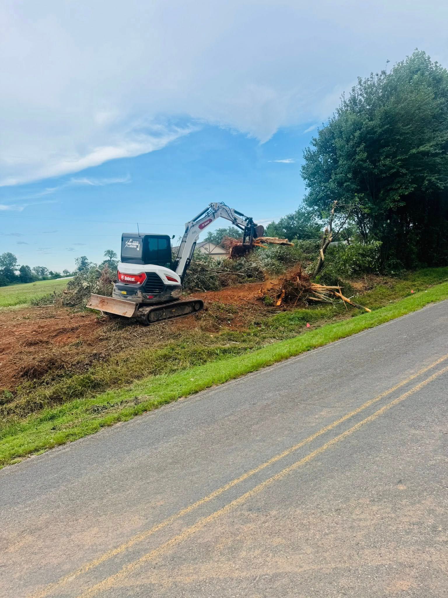 Excavator clearing brush beside a rural road under a cloudy sky