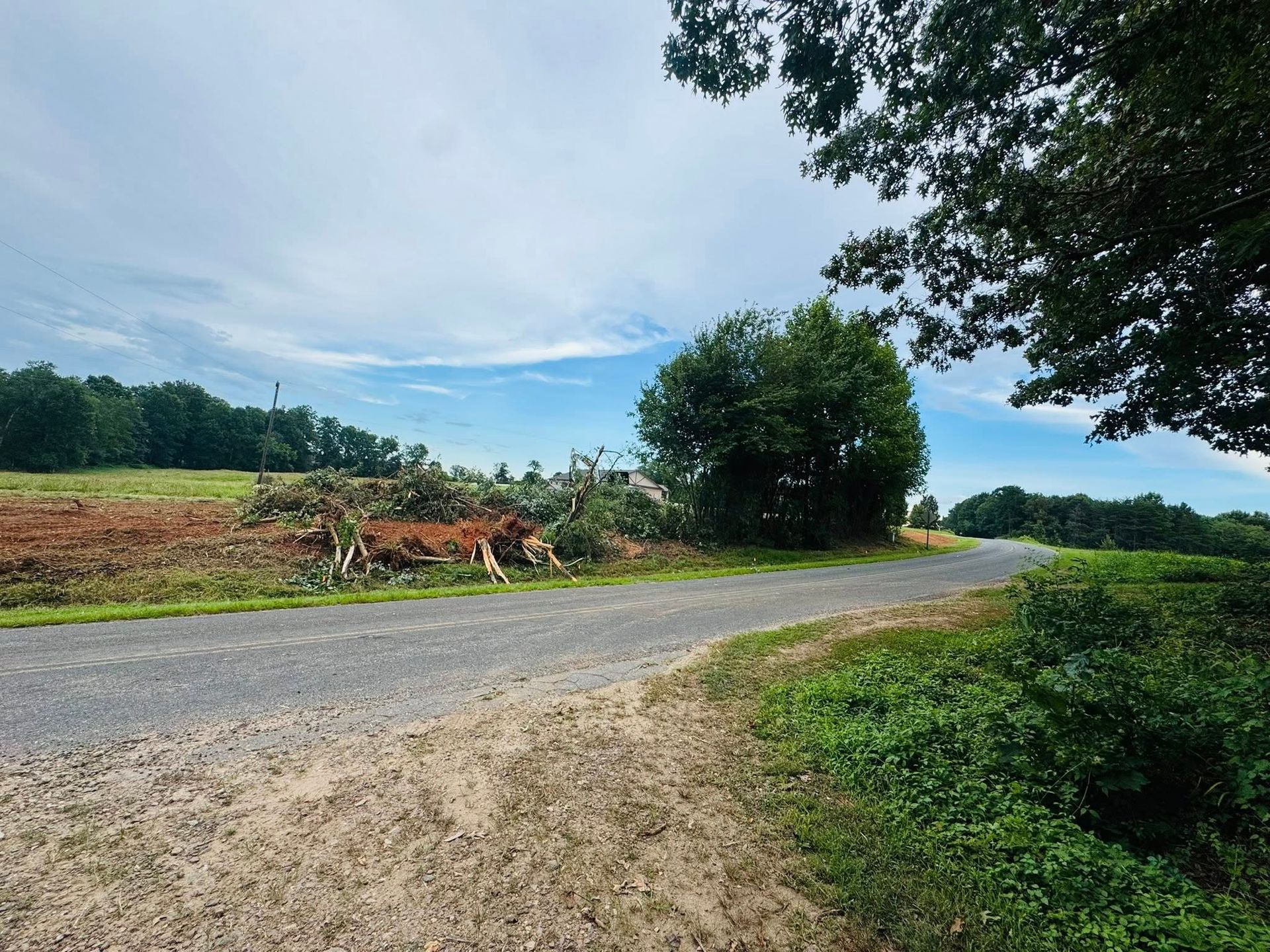 Rural gravel road curving past green fields and trees under a cloudy sky