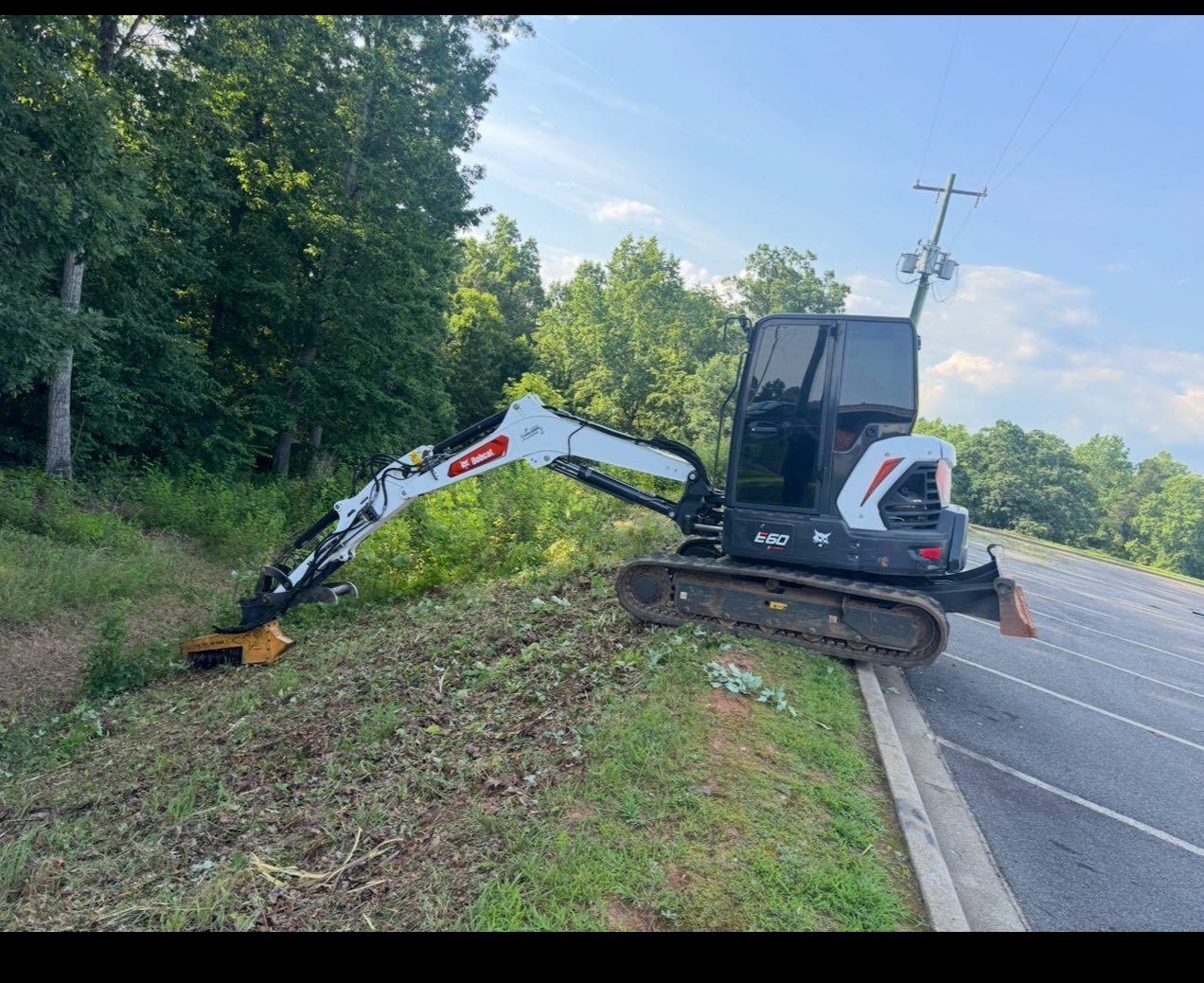 Mini excavator mowing roadside ditch beside a paved road, with trees and utility pole in the background
