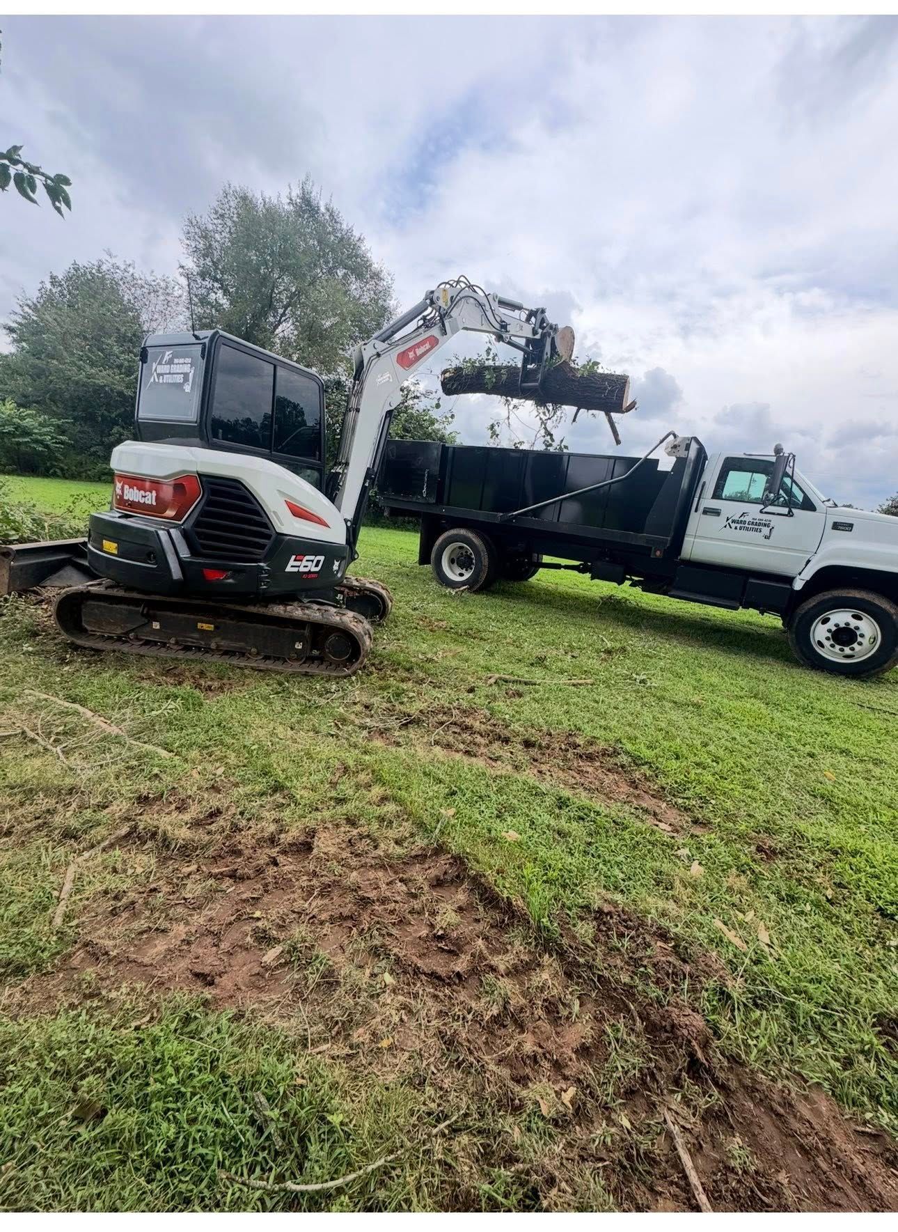 Two construction vehicles on muddy grass, one lifting equipment with an excavator arm under cloudy sky