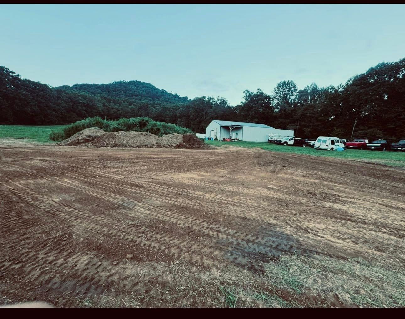 Open field with a white building, parked vehicles, and wooded hills under a pale sky