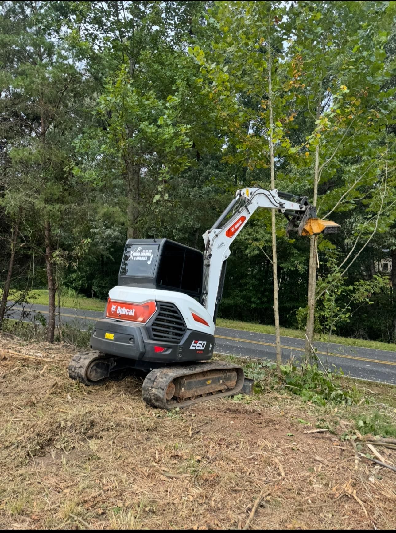 Compact white-and-orange excavator clearing brush beside a road in a wooded area