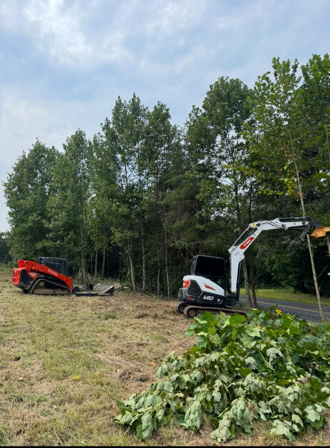 Red excavator and white skid steer clearing brush beside a road and trees