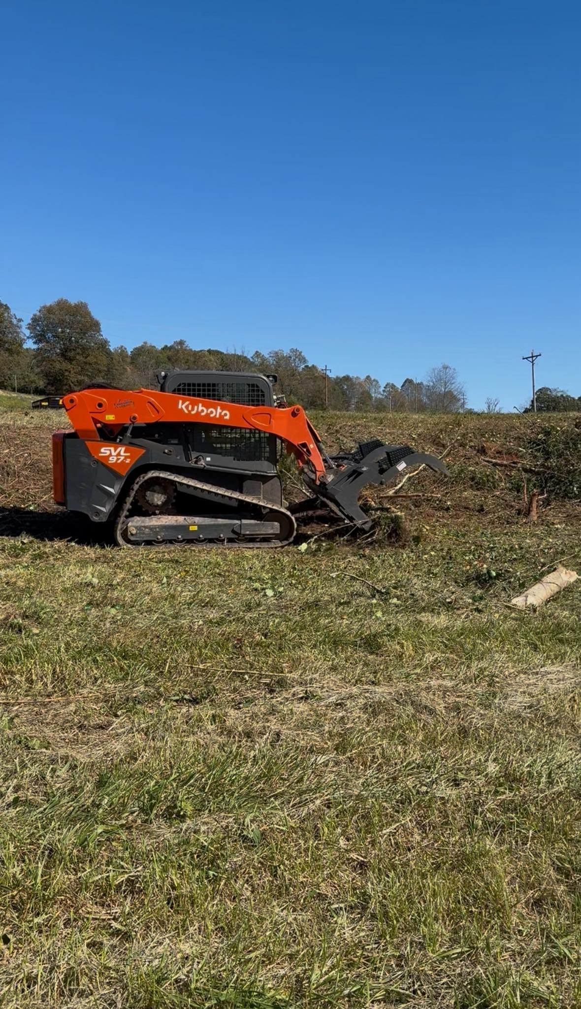 Orange excavator clearing brush in a grassy field under a blue sky.