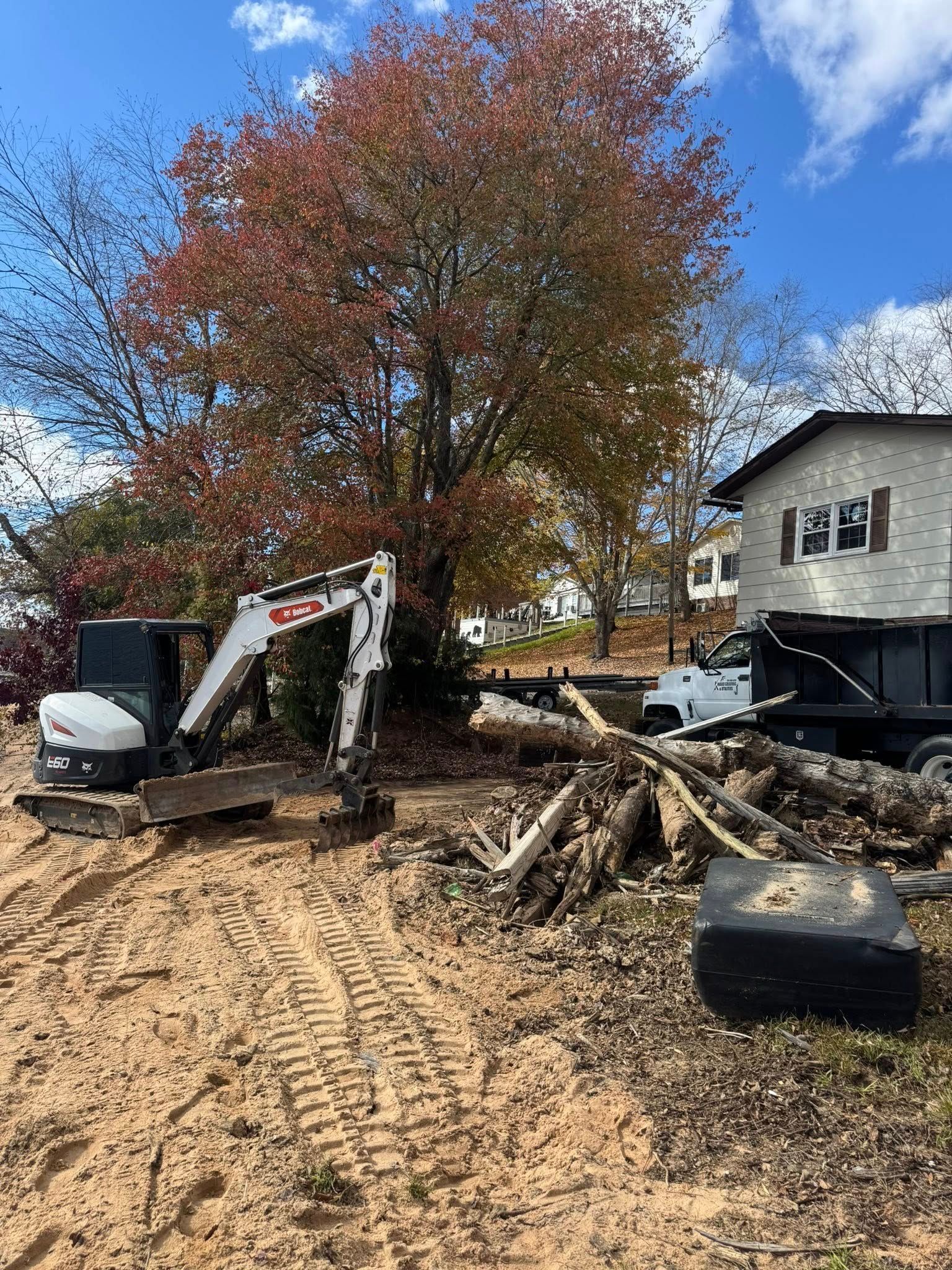 Construction site with excavator, muddy ground, fallen tree limbs, and a house beside autumn trees under a blue sky