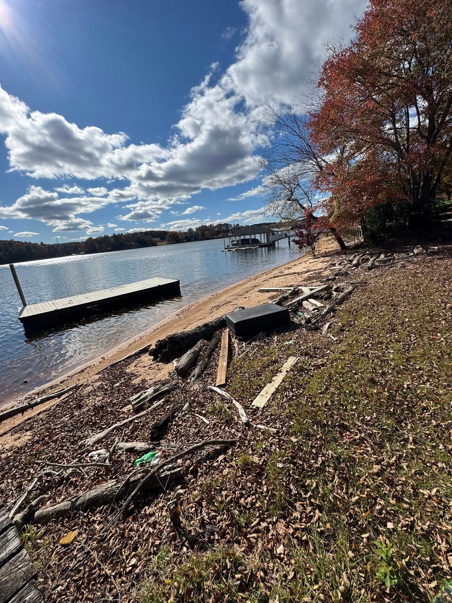 Rocky lakeshore with a dock, calm water, and a red-leaved tree under a partly cloudy sky