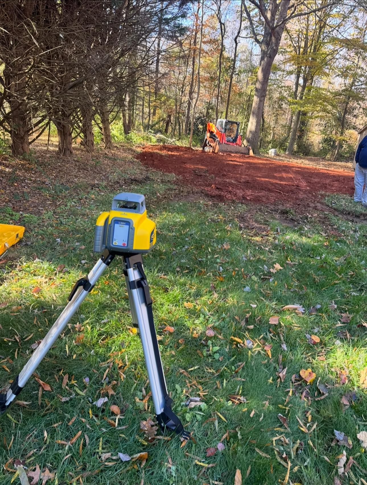 Survey tripod in a wooded clearing with workers in orange vests in the background.