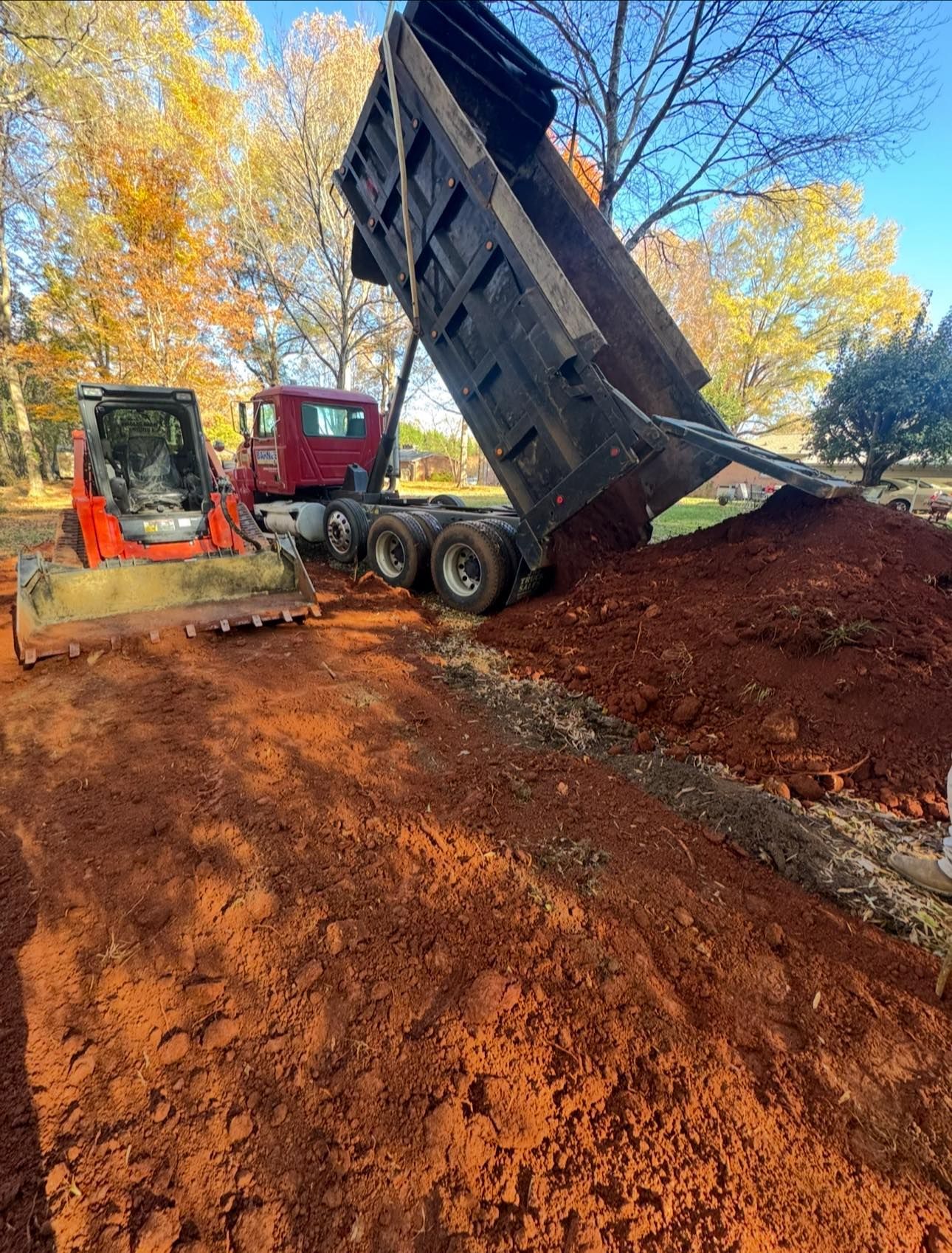Dump truck unloading red dirt beside an excavator on a wooded construction site