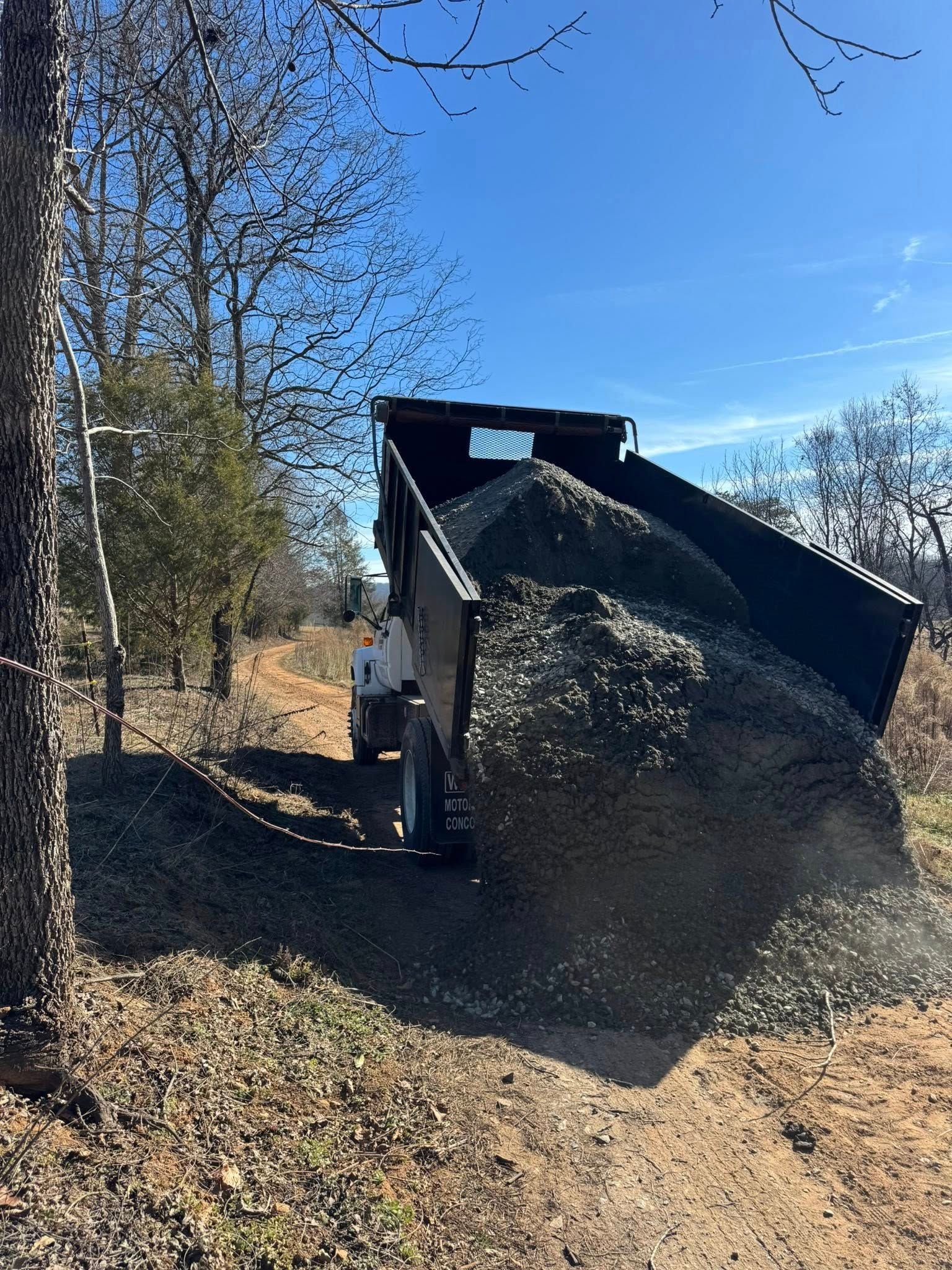 Black dump truck unloading gravel on a sunny dirt road beside trees.