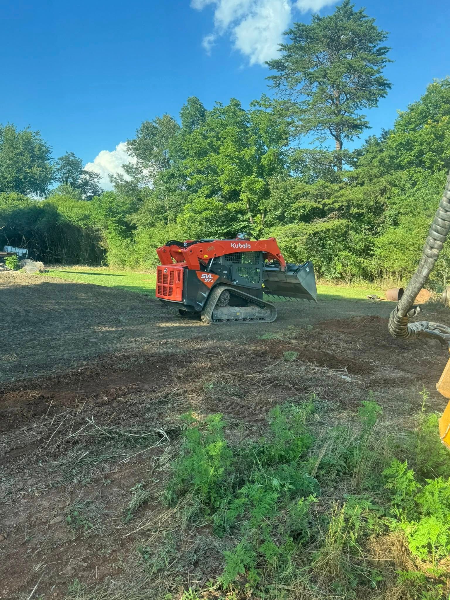Red compact tractor with mower attachment on a gravel clearing beside green trees under a blue sky.