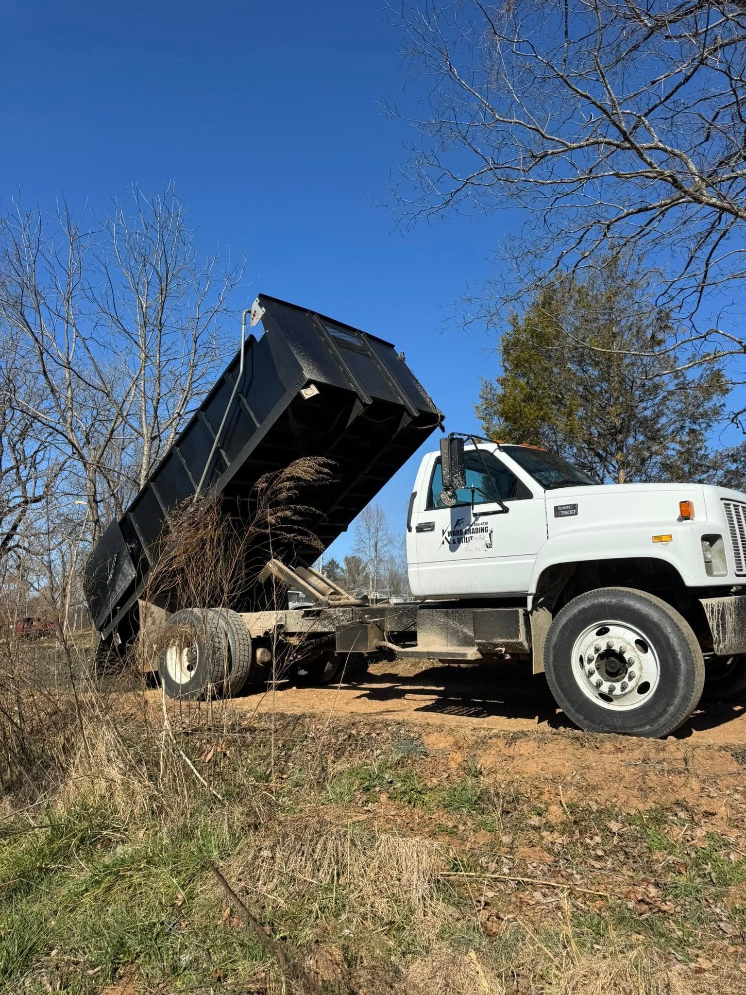 White dump truck raising its black bed in a grassy field under a clear blue sky