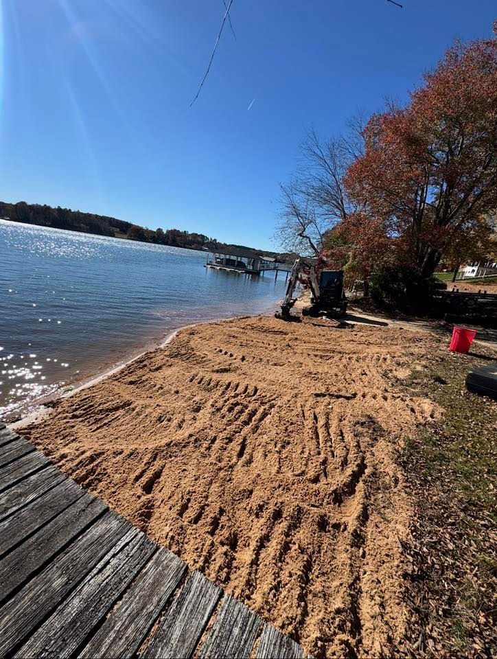 Raked sand beach by a lake under blue sky, with a person near trees and a wooden boardwalk in the foreground
