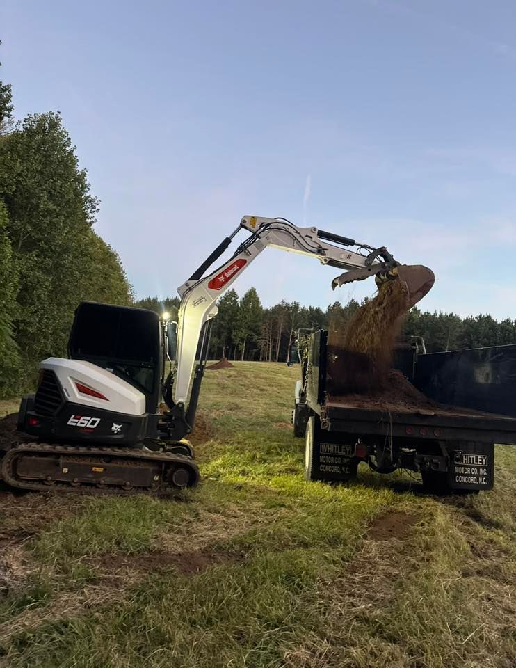 White excavator loading dirt into a dump truck in a grassy field under a blue sky