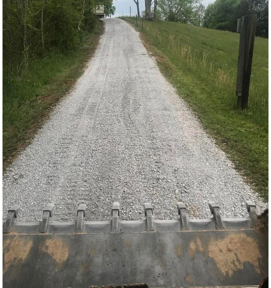 Gravel driveway rising uphill through green fields, viewed from behind a loader bucket.