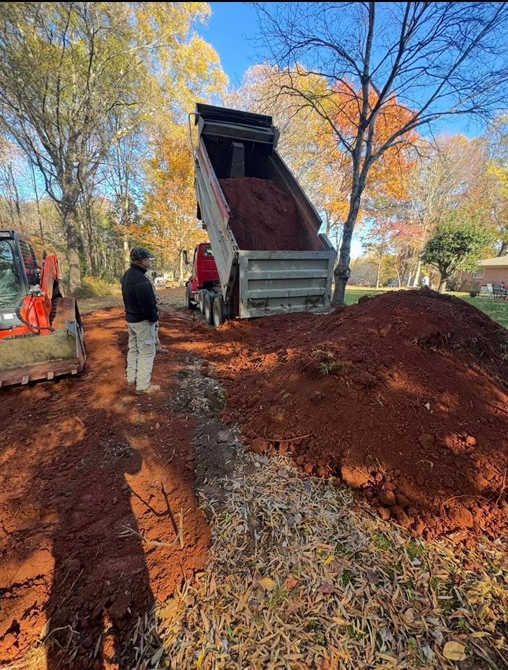 Dump truck unloading red mulch at a park, with workers nearby and autumn trees in the background.