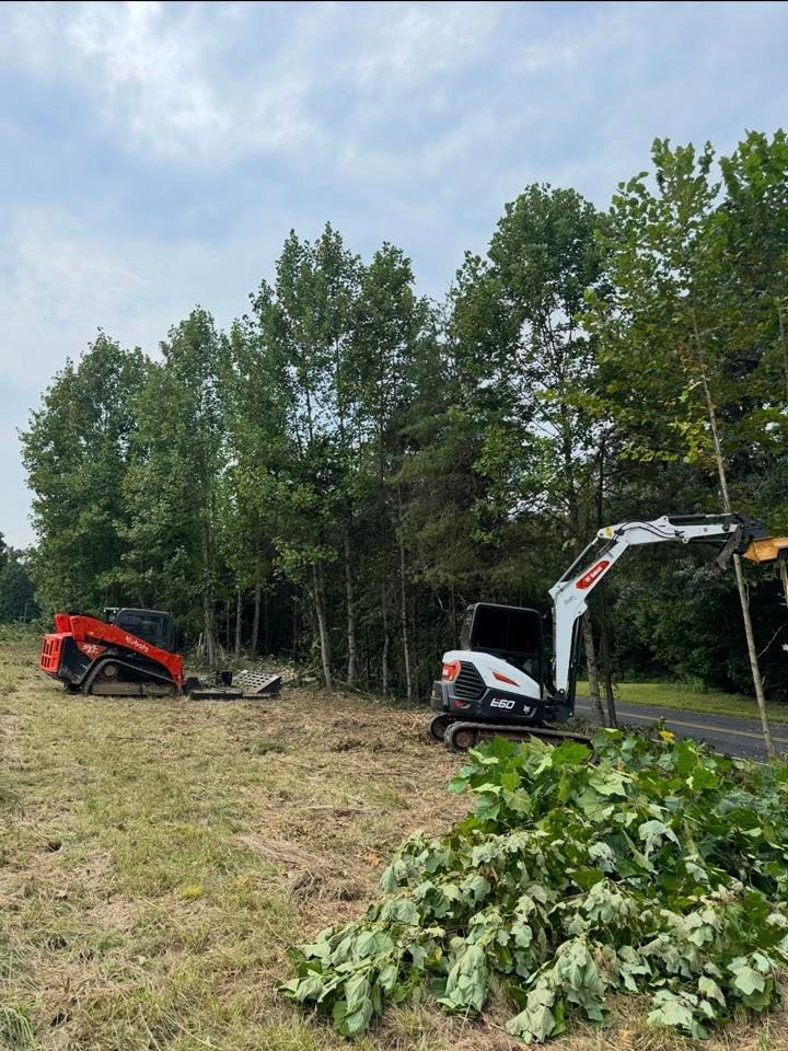 Two utility machines clearing branches beside a wooded roadside, one red and one white with an extended arm